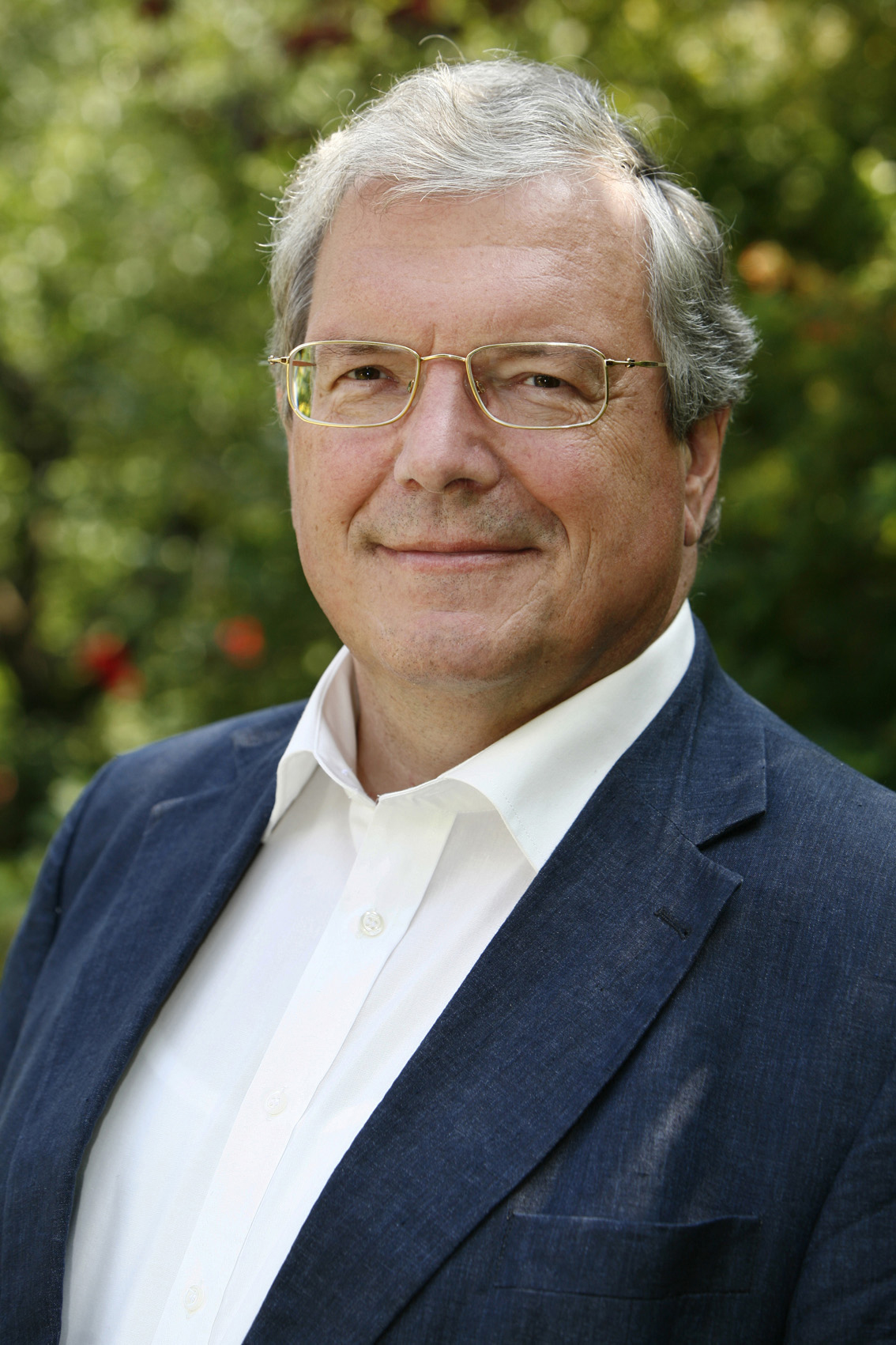 Das Portrait eines Mannes mit weißen Haaren und Brille in einem weißen Hemd und einer blauen Anzugjacke.